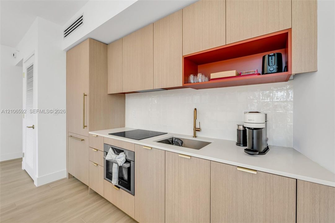 Flat-panel wood-finish cabinetry, white solid-surface countertop, integrated electric cooktop, stainless steel under-mount sink with brass-toned faucet, and gloss-finish wall tiling