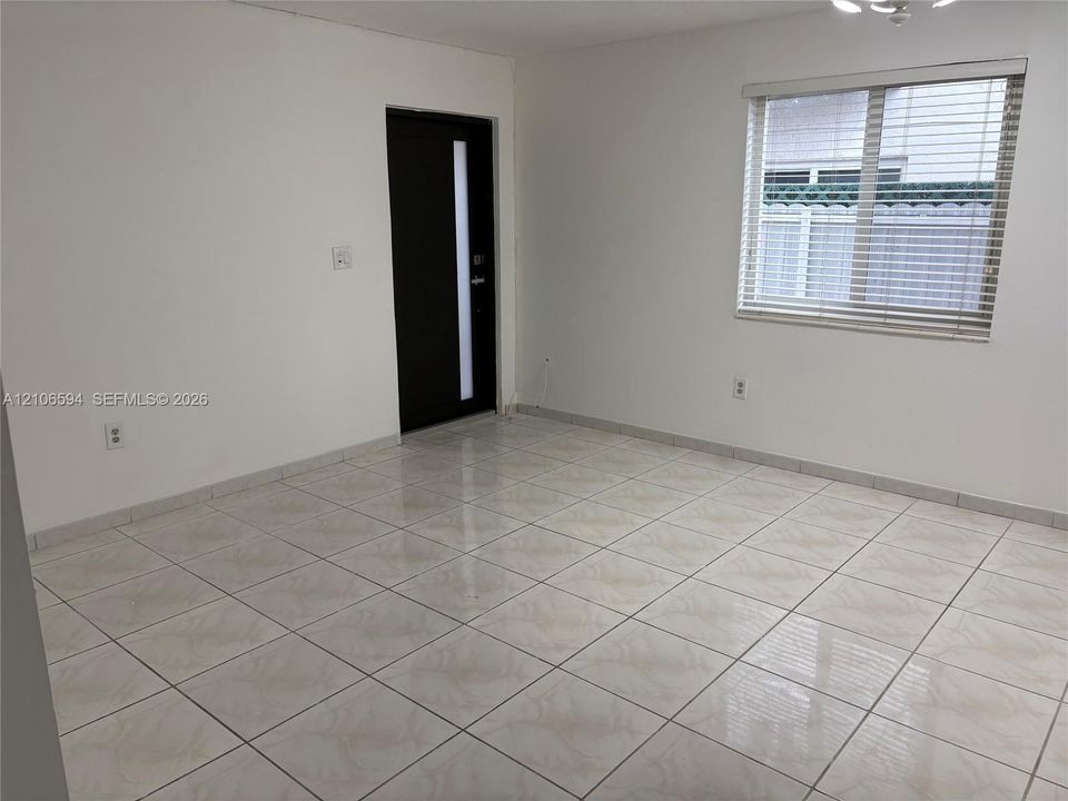 Neutral-toned ceramic tile flooring, solid-surface white walls, window with integrated horizontal blinds, and a modern single-panel entry door featuring a vertical glass inlay