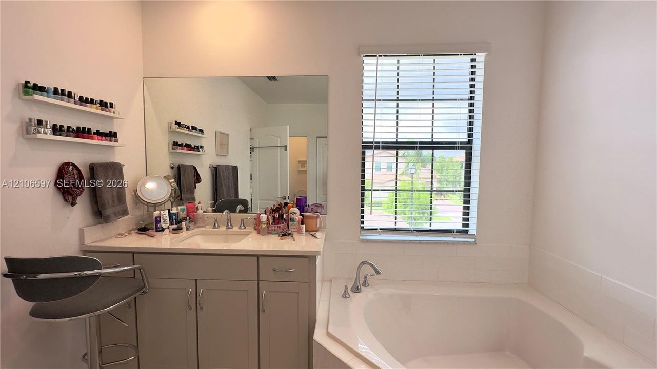Bathroom featuring a vanity with light-colored cabinetry, a stone-composite countertop with an integrated basin, a large wall-mounted mirror, a jetted soaking tub with deck-mounted hardware, and a window with horizontal blinds
