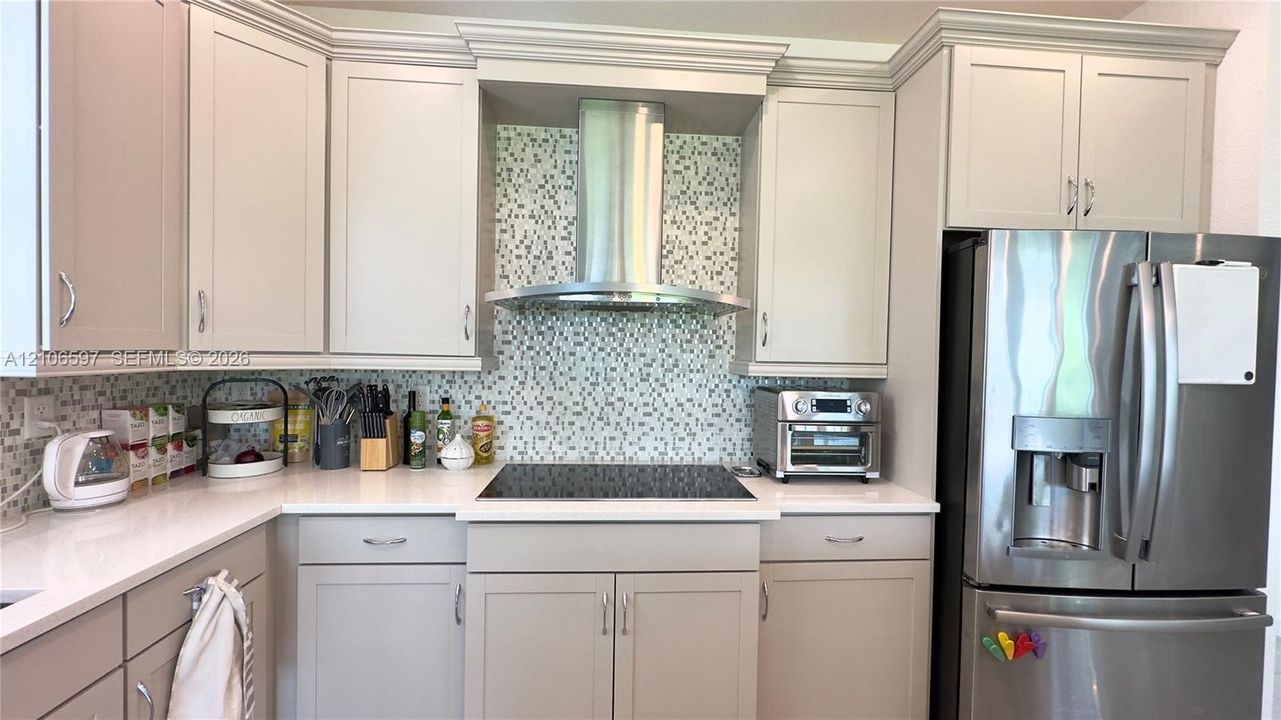 Kitchen featuring neutral-toned cabinetry, solid surface countertops, a mosaic tile backsplash, a stainless steel chimney-style range hood, and a built-in stainless steel refrigerator