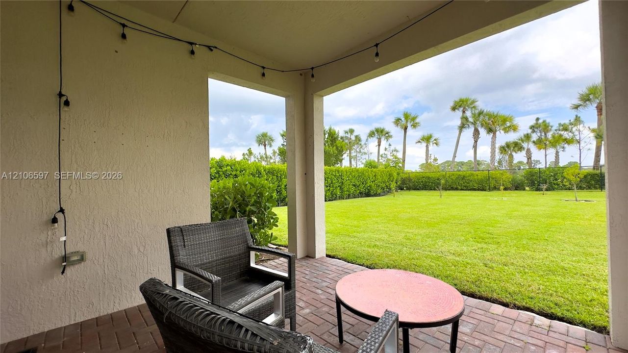 Covered patio featuring brick pavers, textured stucco walls, and an expansive lawn with mature perimeter shrubbery and palm trees