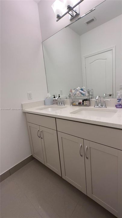 Upstairs guest bathroom Dual-sink vanity featuring light-toned cabinetry, light-toned stone-finish countertop, undermount sinks, and a large-format frameless mirror with mounted lighting fixture