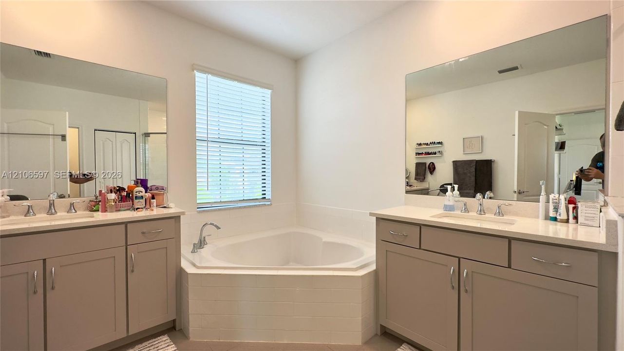 Dual vanities featuring shaker-style cabinetry, solid-surface countertops, expansive wall-mounted mirrors, and an integrated corner soaking tub with a tiled surround