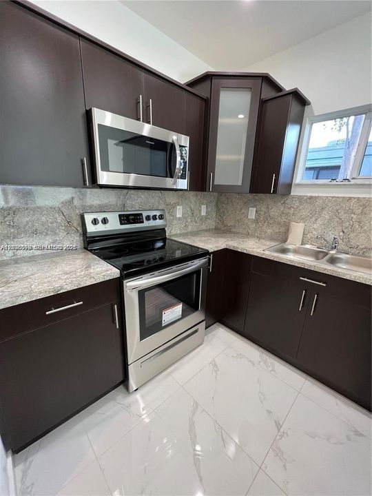 Dark-finish cabinetry with brushed metal hardware, stone-style countertops, stainless steel appliance suite, glass-paneled upper cabinet, and large-format tile flooring