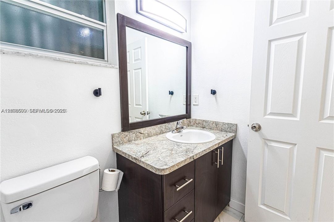 Dark wood cabinetry with brushed nickel hardware, stone-finish countertop, integrated oval basin, framed vanity mirror, and horizontal wall-mounted light fixture