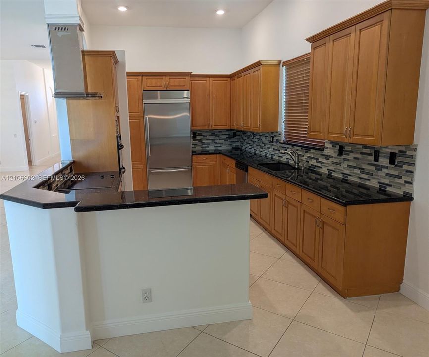 Kitchen featuring wood cabinetry, dark stone countertops, stainless steel refrigerator, tile backsplash, and a stainless steel wall-mounted range hood