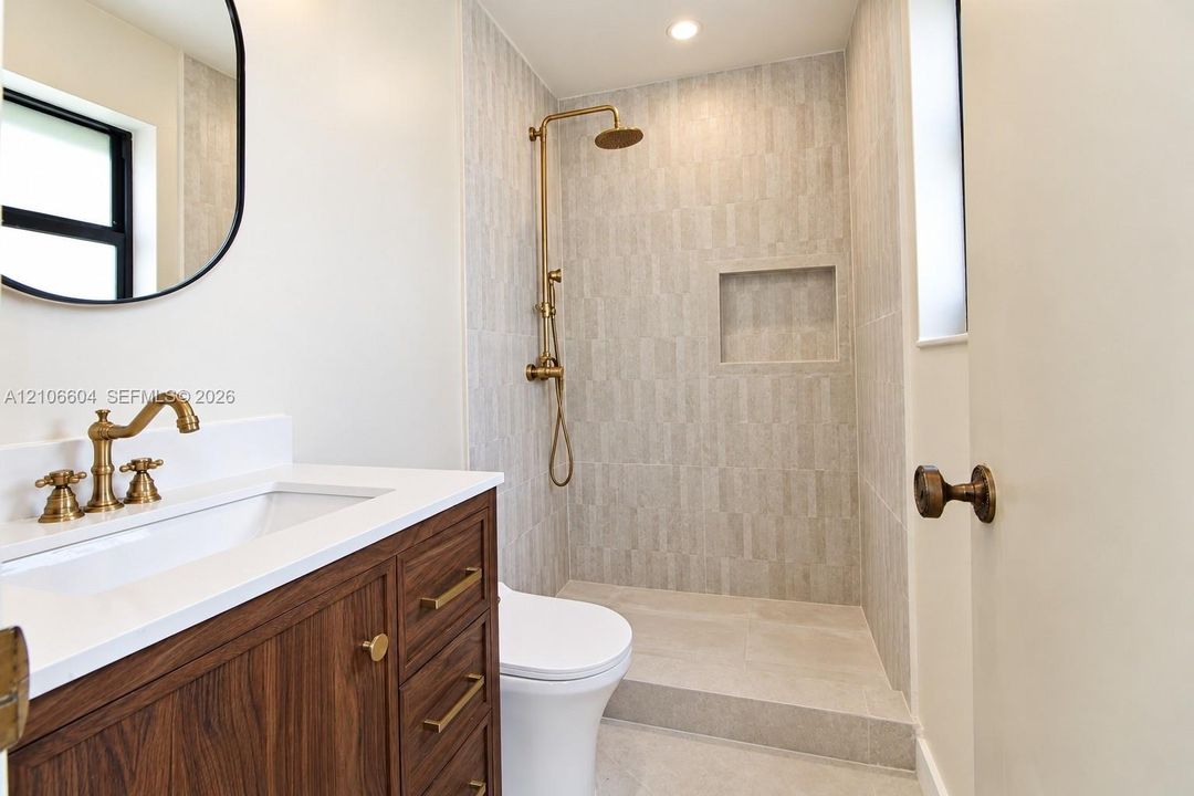 Wood-finish vanity with white countertop, brass-toned fixtures, walk-in shower with vertical tile surround and built-in niche, and brass hardware accents