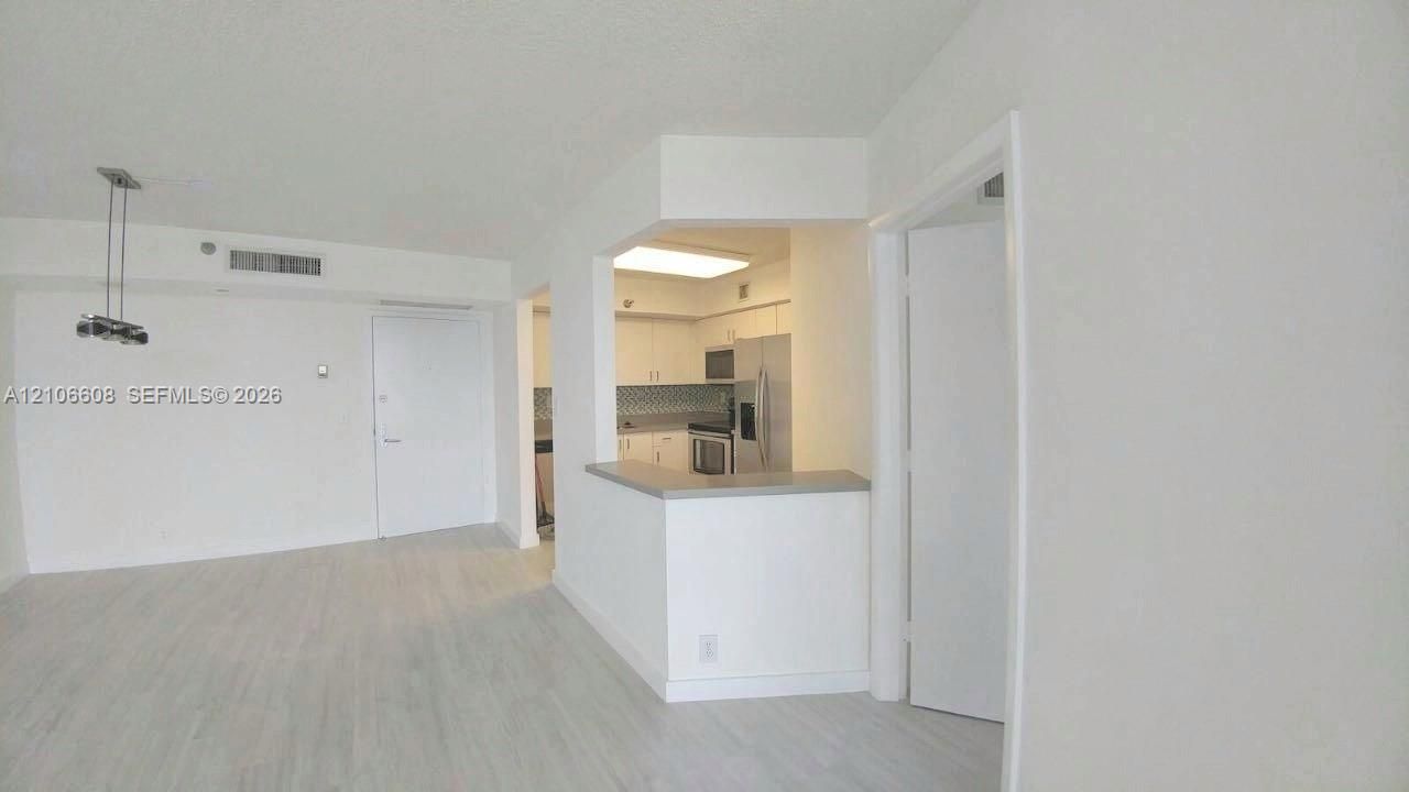 Open-concept interior featuring light-toned wood-finish flooring, a kitchen pass-through counter, neutral cabinetry, patterned tile backsplash, and a modern multi-light pendant fixture