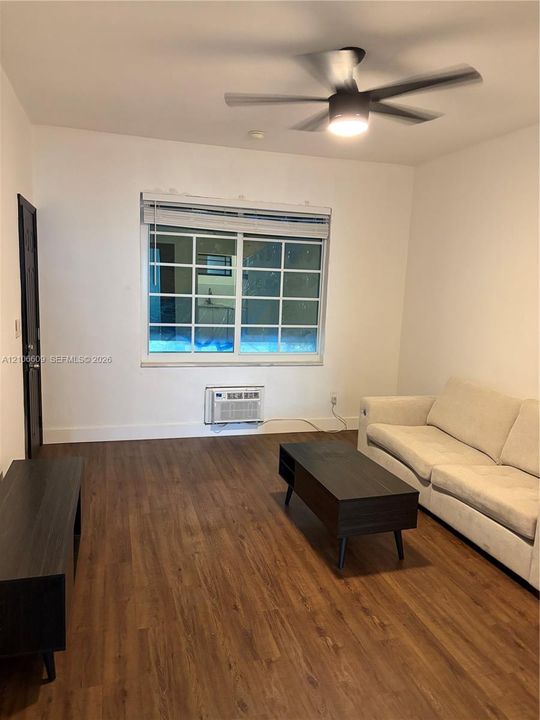 Wood-finish flooring, multi-pane window with horizontal blinds, integrated wall-mounted air conditioning unit, and a central ceiling fan with light fixture
