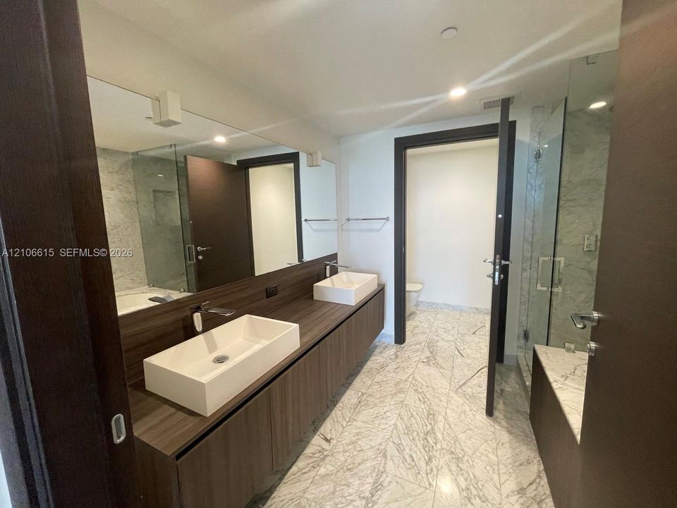 Floating wood-finish vanity with dual rectangular vessel sinks, stone-patterned tile flooring, expansive wall-mounted mirror, and glass-enclosed shower with stone-patterned wall surround