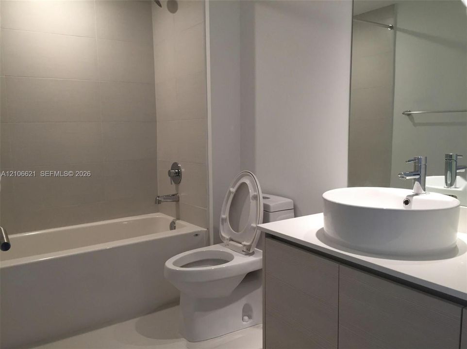 Bathroom featuring a vanity with a rectangular vessel sink, white solid surface countertop, wood-finish cabinetry, a bathtub with full-height tile surround, and a large-format mirror