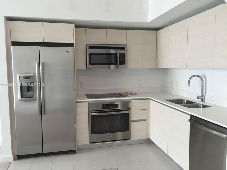 Kitchen featuring light wood-finish cabinetry, white solid-surface countertops, and a suite of stainless steel appliances, including an integrated refrigerator, built-in microwave, range, and dishwasher with a gooseneck faucet