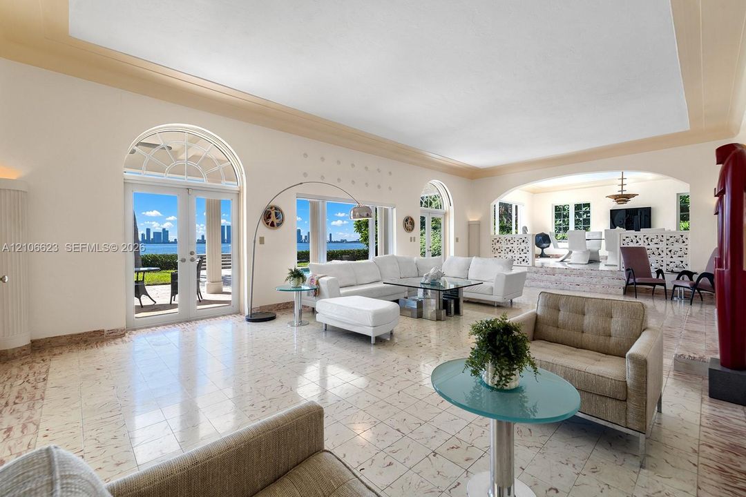 Open-concept interior featuring light-toned stone flooring, arched glass double doors, elevated platform area with decorative latticework, and recessed tray ceiling detailing