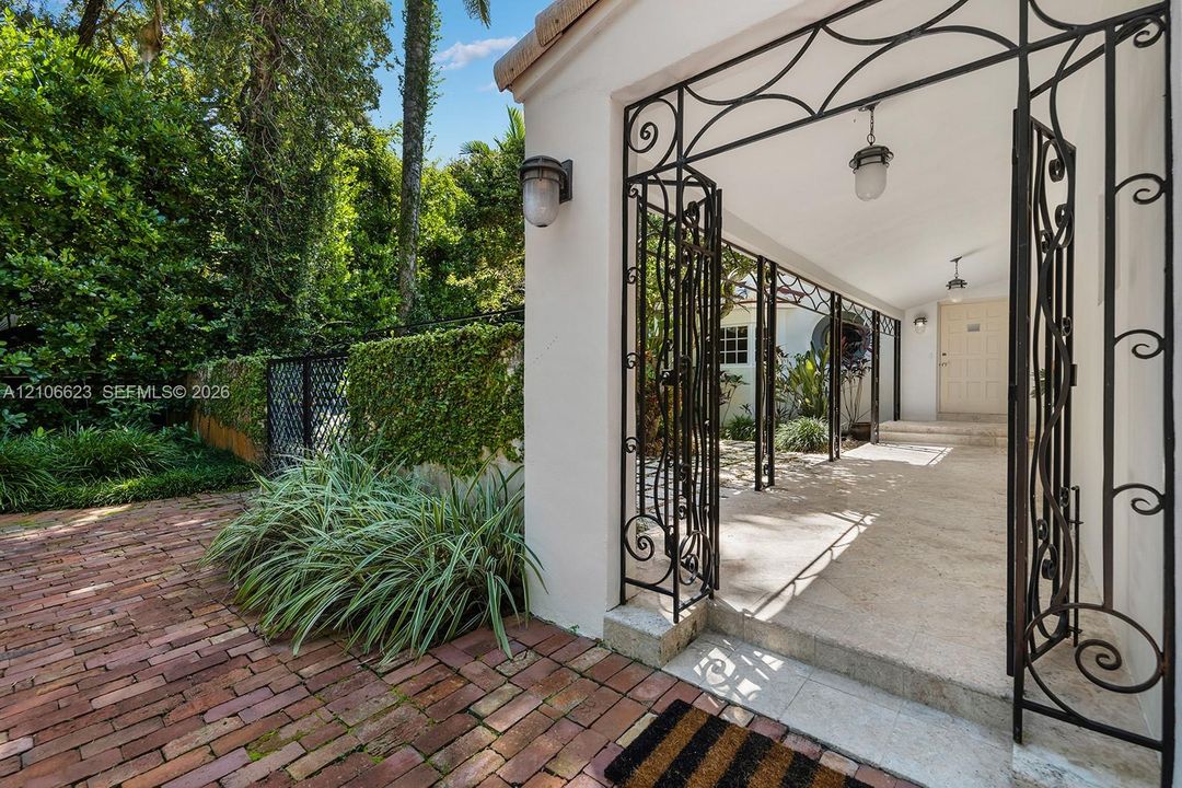 Covered entry featuring decorative wrought-iron gates, exterior wall-mounted lighting, stone flooring, and a brick-paved patio