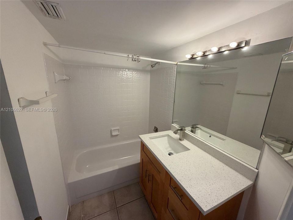 Bathroom featuring wood-finish vanity cabinetry, a stone-flecked composite countertop with an integrated basin, a large wall-mounted vanity mirror, a multi-bulb light fixture, and a tiled shower-tub combination