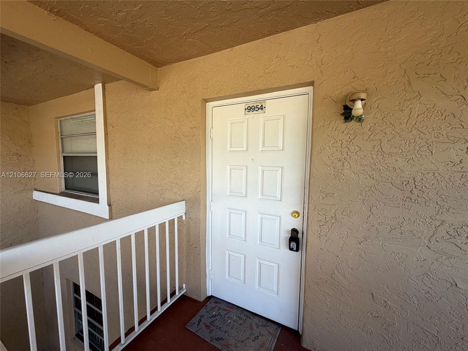 Exterior entryway featuring a white paneled door, textured stucco wall finish, metal railing, and wall-mounted exterior lighting