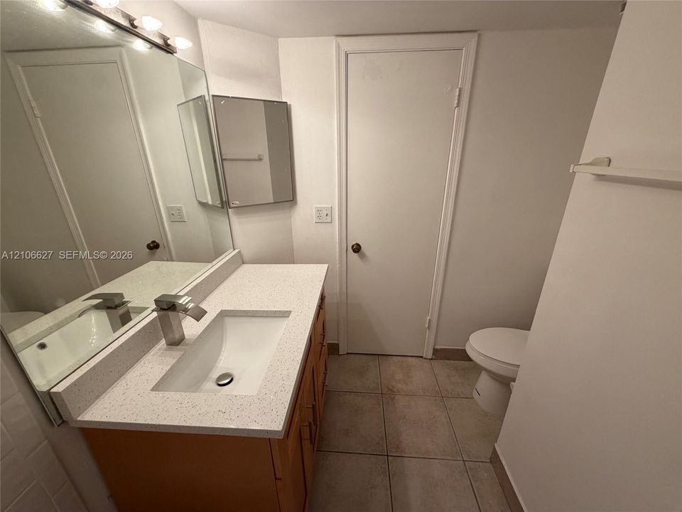 Vanity featuring a stone-composite countertop with an integrated rectangular sink and modern metal faucet, complemented by a large wall-mounted mirror, ceramic tile flooring, and neutral wall cabinetry