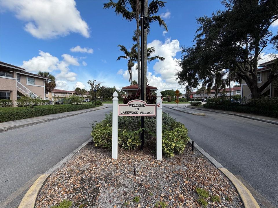 Lakewood Village community signage situated within a landscaped traffic island featuring stone mulch, manicured shrubbery, and mature palm trees
