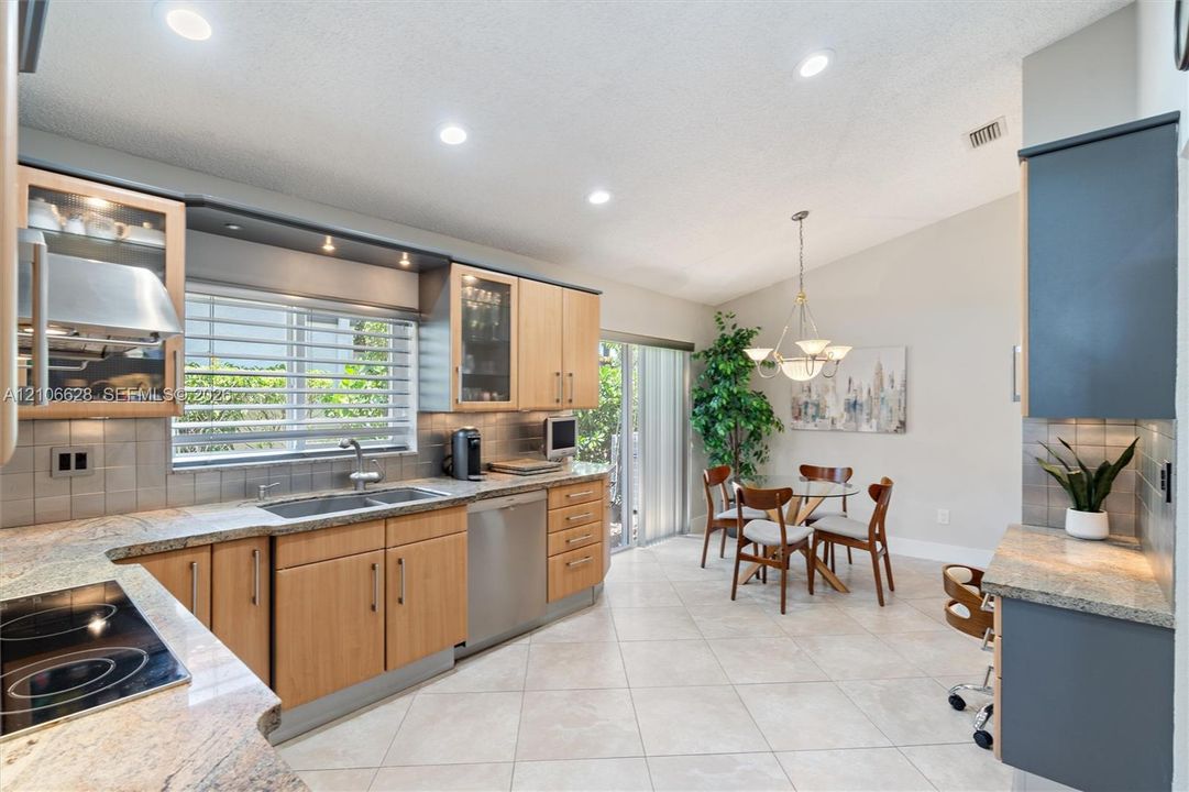 Kitchen featuring light-finish wood cabinetry, integrated stainless steel appliances, neutral stone-finish countertops, tile backsplash, and ceramic tile flooring