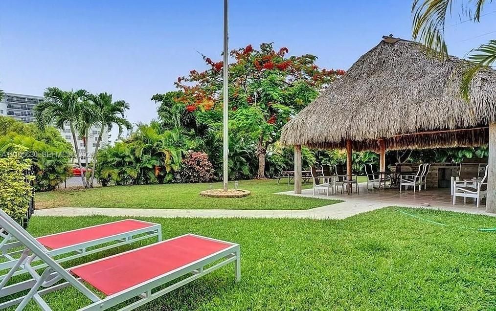 Expansive backyard featuring a thatched-roof palapa, stone-finish outdoor kitchen, manicured lawn, and mature tropical landscaping