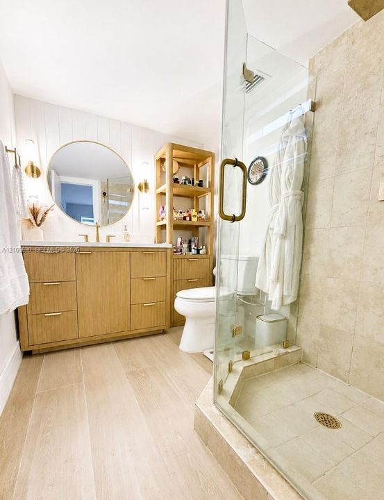 Bathroom featuring wood-finish cabinetry with brass-tone hardware, a circular wall-mounted mirror, light-toned wood-finish flooring, and a glass-enclosed shower with stone-tile surround