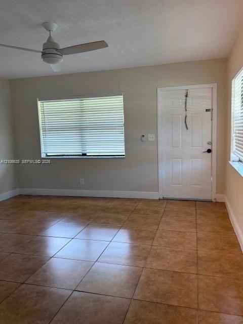Neutral-toned interior featuring large-format tile flooring, a white six-panel entry door, horizontal window blinds, and a white ceiling fan