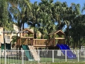 Outdoor recreation area featuring a wooden play structure with integrated slides and stairs, enclosed by white metal fencing and surrounded by mature palm trees