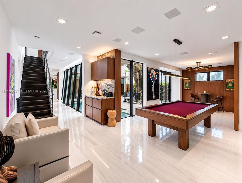 Polished stone flooring, wood-panelled accent walls, integrated bar cabinetry with stone backsplash, staircase with open risers, and floor-to-ceiling glass paneling