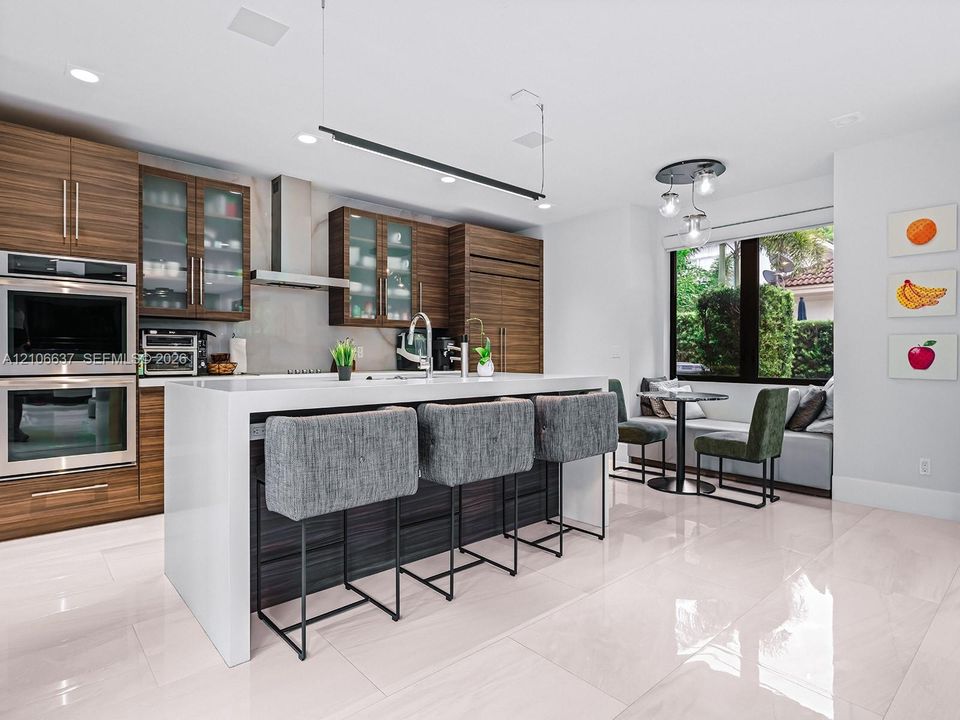 Modern kitchen featuring horizontal-grain wood cabinetry, a central island with a waterfall-edge countertop, built-in double wall ovens, recessed lighting, and large-format tile flooring