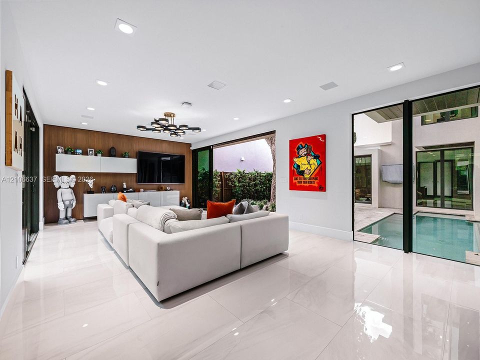 Large-format tile flooring, wood-panel accent wall with integrated built-in cabinetry, recessed ceiling lighting, statement multi-arm chandelier, and floor-to-ceiling glass doors opening to an internal pool area
