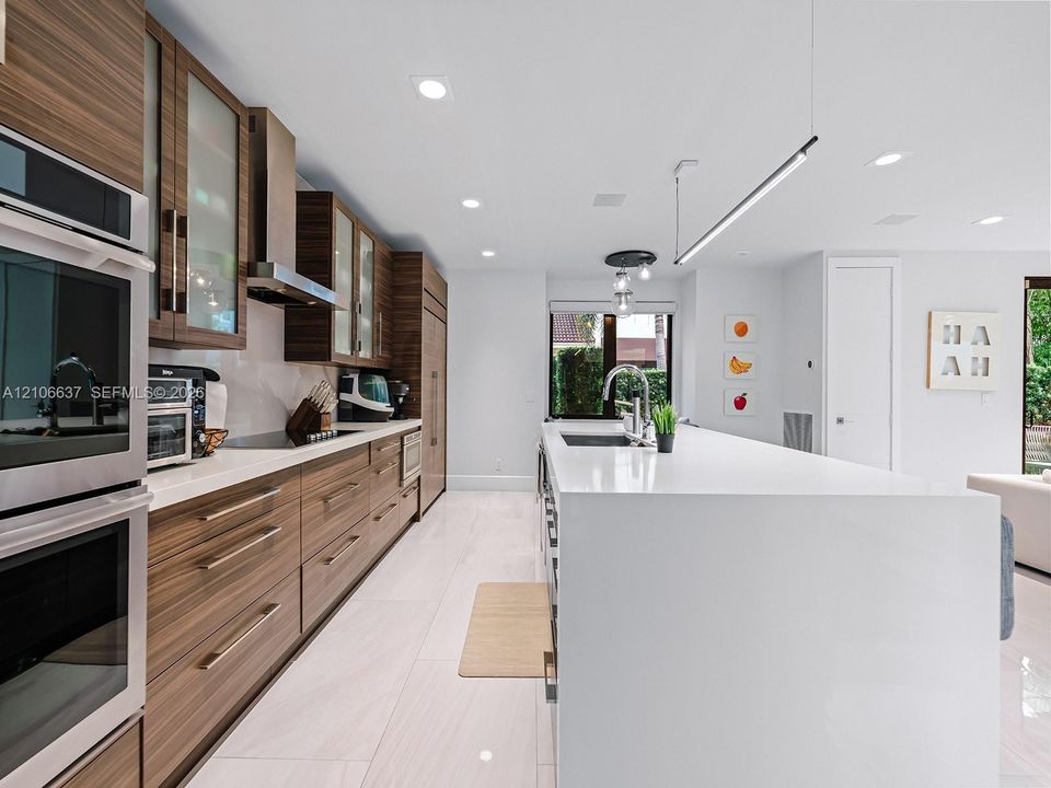Contemporary kitchen featuring horizontal-grain wood cabinetry, expansive solid-surface island, stainless steel built-in double ovens, integrated stainless steel range hood, and large-format porcelain tile flooring