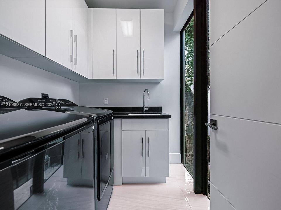 Glossy white cabinetry with vertical pulls, solid-surface dark countertop, undermount sink with gooseneck faucet, light-toned polished flooring, and full-height glass paneled door