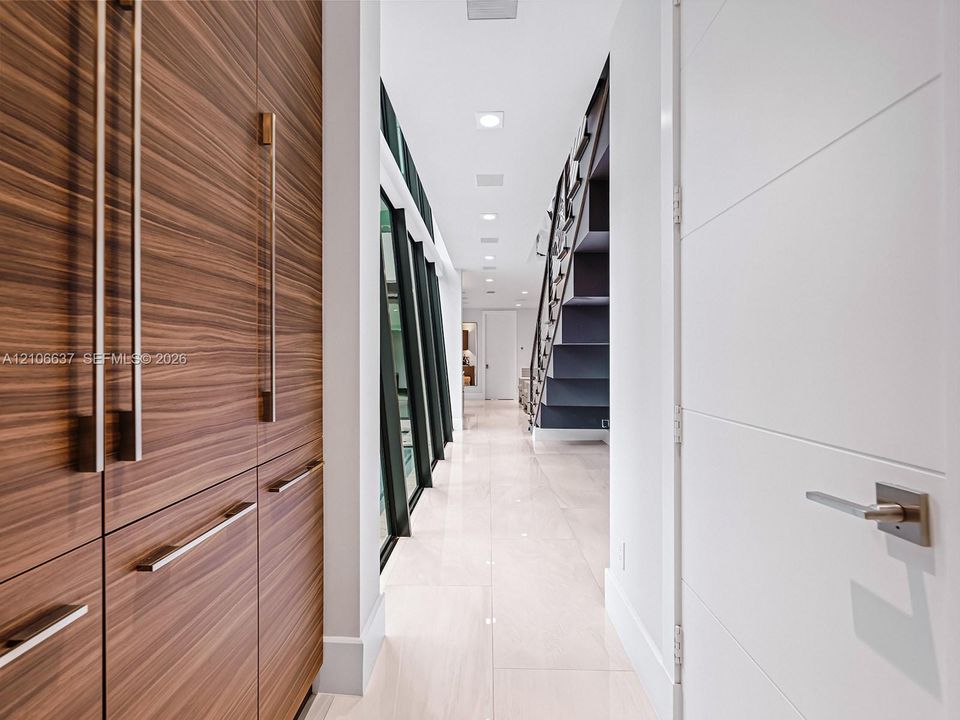 Hallway featuring wood-finish cabinetry with vertical hardware, large-format tile flooring, recessed ceiling lighting, and a dark-toned staircase with metal handrails