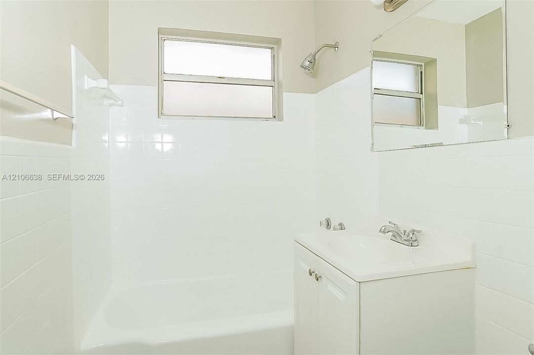 Bathroom featuring white tile wall surround, integrated tub and shower fixture, rectangular window, vanity with solid surface countertop, and wall-mounted mirror