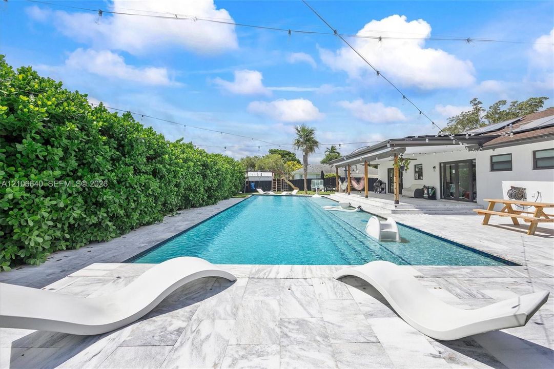 Expansive outdoor living area featuring a rectangular swimming pool, large patio with light-toned pavers, and a covered patio structure