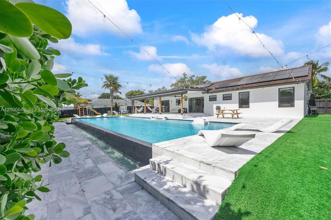 Expansive outdoor area featuring a swimming pool with a dark tile edge, a large patio with light-colored tile, and a covered patio area
