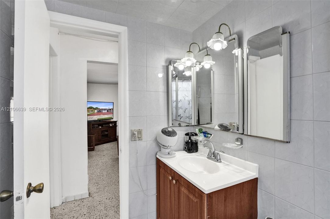 Vanity with a solid surface countertop, integrated sink, wooden cabinetry, wall-mounted mirror, and multi-bulb light fixture, finished with full-height tile wall surfacing