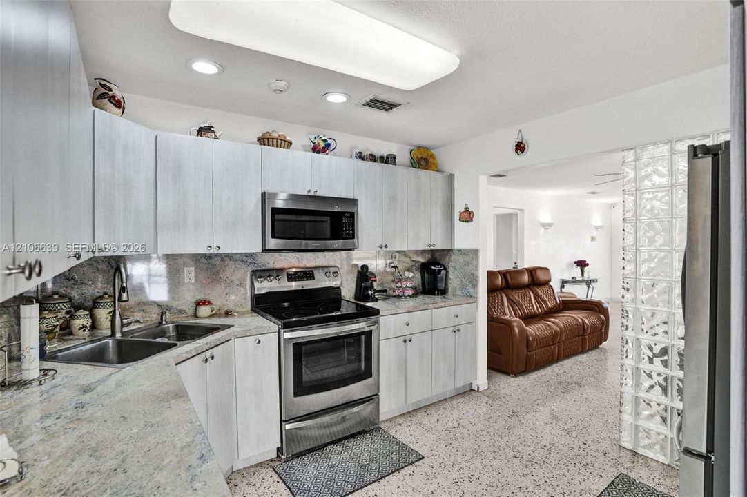 Kitchen featuring light-finish cabinetry, stone countertops, stainless steel appliances, a double-basin stainless steel sink, and a decorative glass block partition
