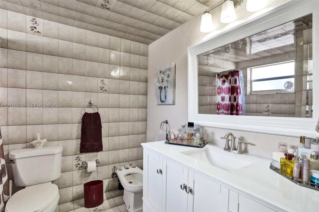 White vanity with a smooth integrated countertop and dual-handle faucet, expansive wall-mounted mirror, three-bulb vanity light fixture, bidet, and floor-to-ceiling decorative tiled wall finishes