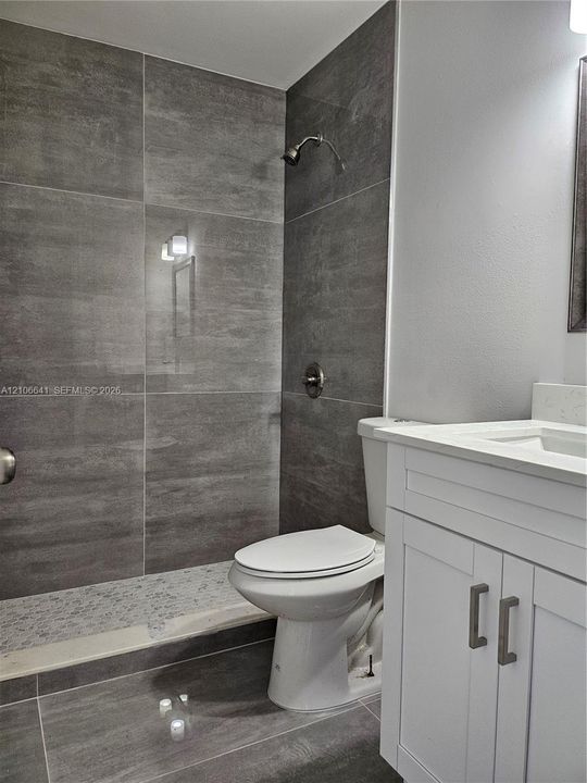 Bathroom featuring large-format gray tile shower surround, integrated pebble-stone shower floor, white vanity cabinet with stone-finish countertop, and wall-mounted brushed metal hardware
