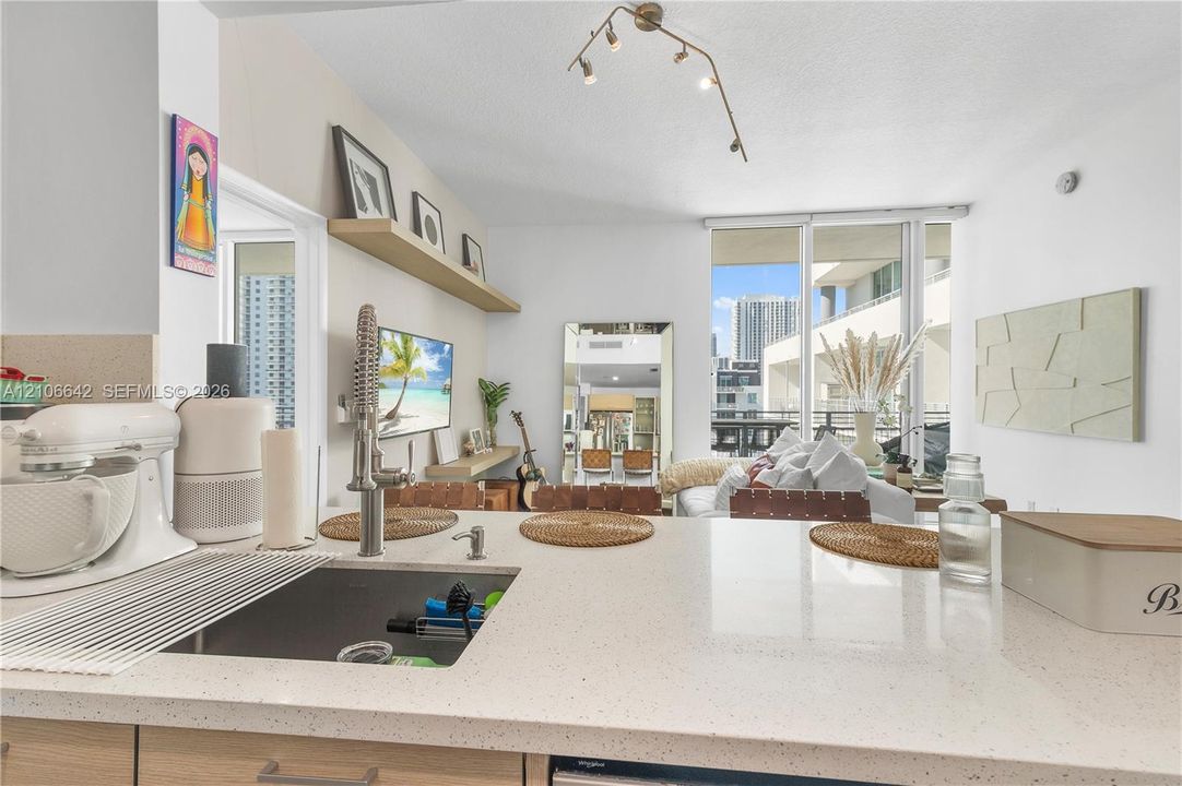 Kitchen featuring quartz countertops, an undermount stainless steel sink with a high-arch pull-down faucet, wood-finish cabinetry, built-in wall shelving, and floor-to-ceiling glass paneling