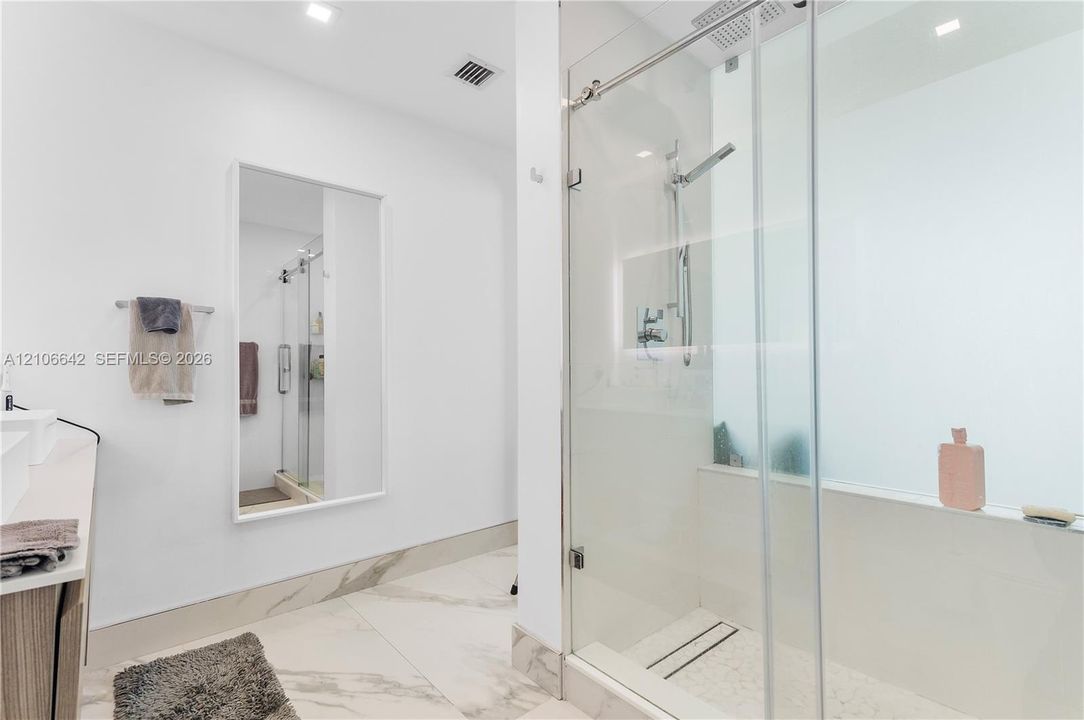 Walk-in shower with a glass enclosure, wall-mounted rainfall showerhead, stone-finish tile flooring, recessed ceiling lighting, and a large wall-mounted mirror
