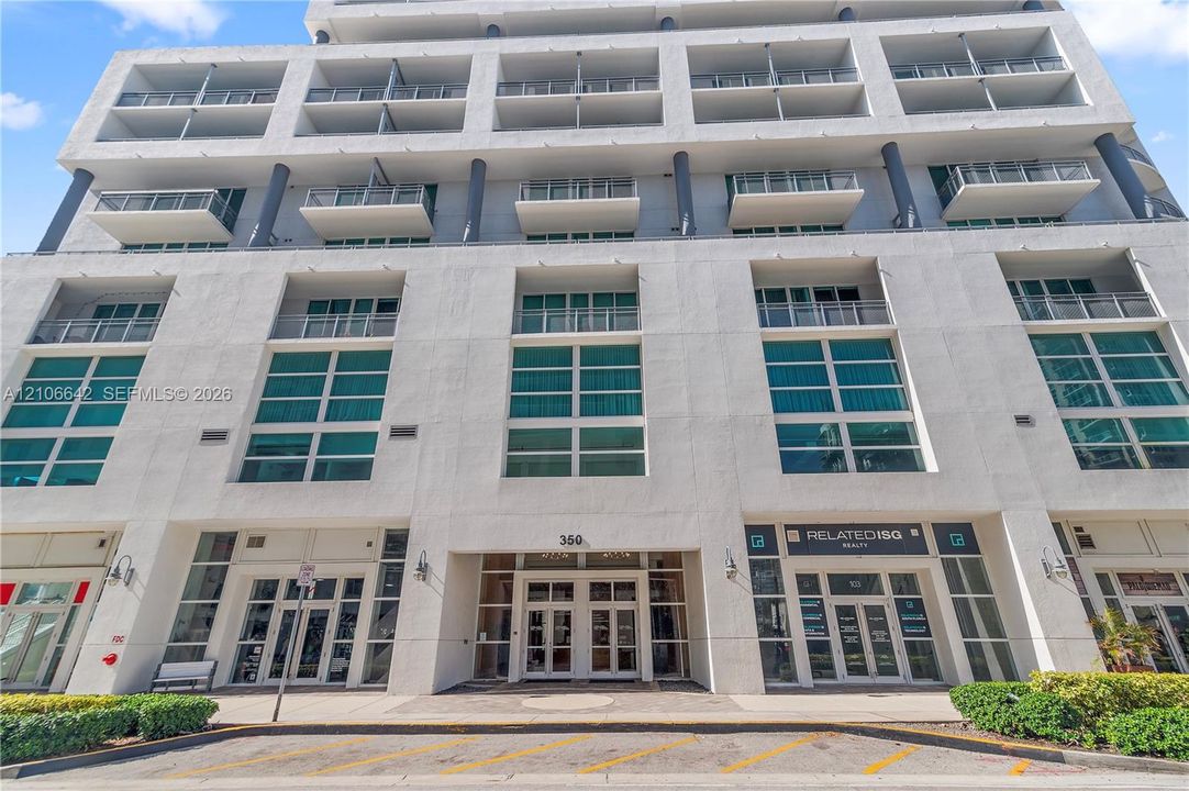 Multi-story concrete exterior featuring floor-to-ceiling glass windows, private balconies with metal railings, and a recessed ground-level entryway