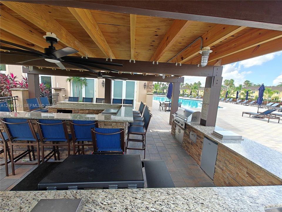 Outdoor kitchen featuring a stone-topped counter, covered timber-frame pergola, expansive paver patio, metal perimeter fencing, and a pool