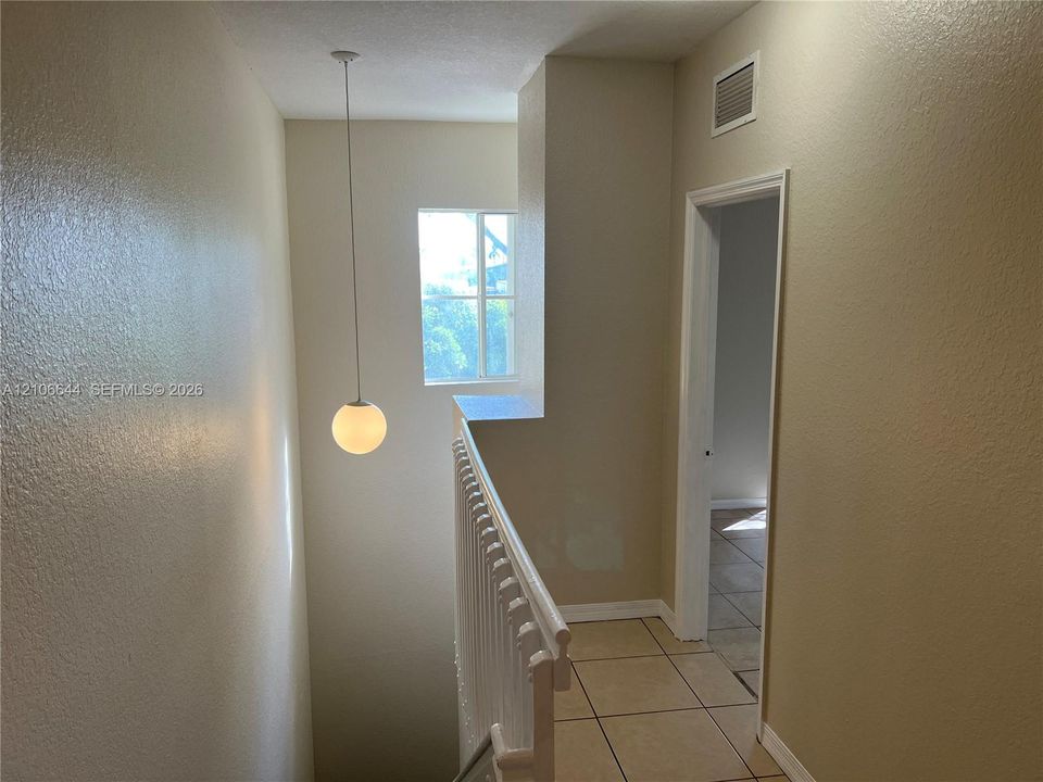 Neutral-toned interior hall featuring white ceramic tile flooring, a spherical pendant light fixture, a square window, and a white balustrade railing
