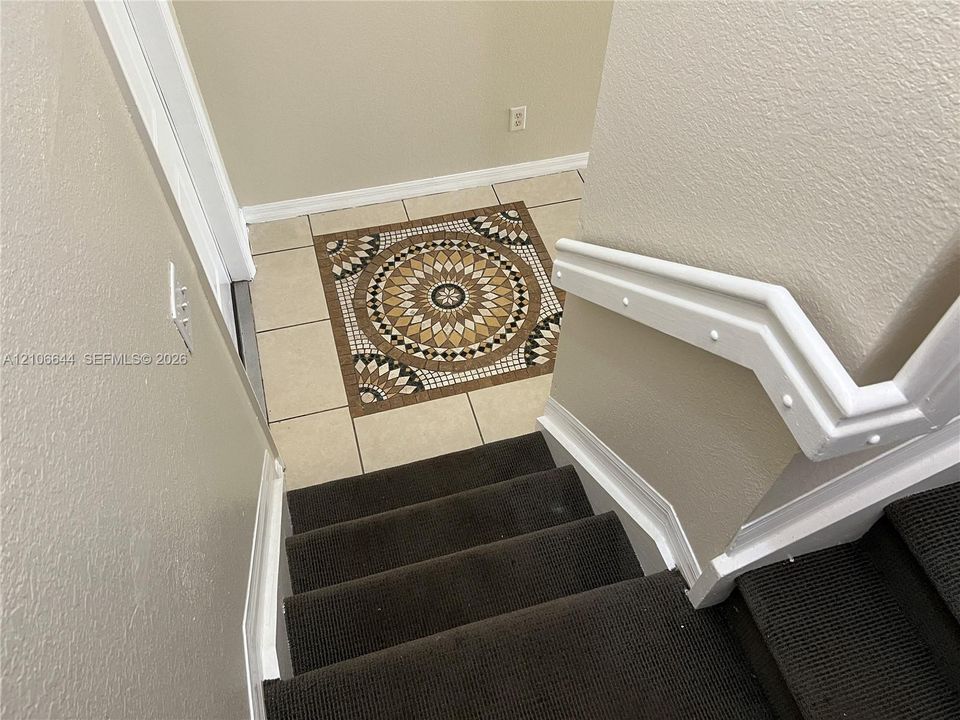 Staircase featuring dark carpeting, white handrails, and neutral wall tones, punctuated by a tiled floor with a decorative mosaic medallion inlay