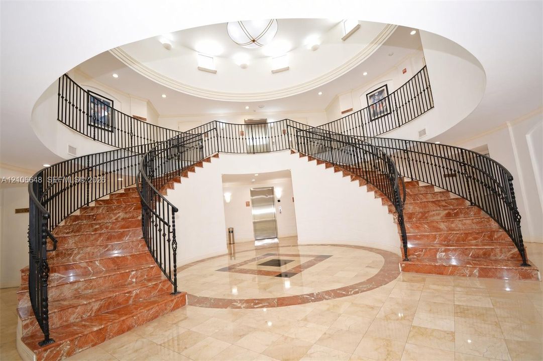 Grand dual staircase featuring stone-finish treads, decorative metal railings, a circular tray ceiling, tiled flooring with inlaid detailing, and a stainless steel elevator