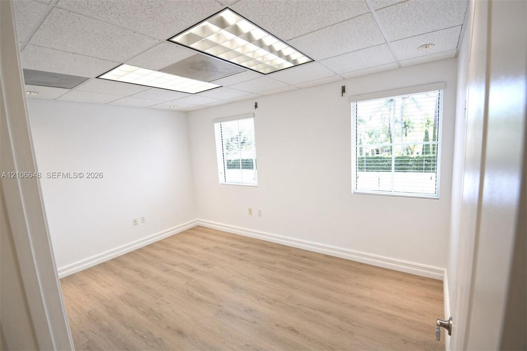 Neutral-toned room featuring wood-finish flooring, two windows with horizontal blinds, and a suspended drop ceiling with recessed lighting panels