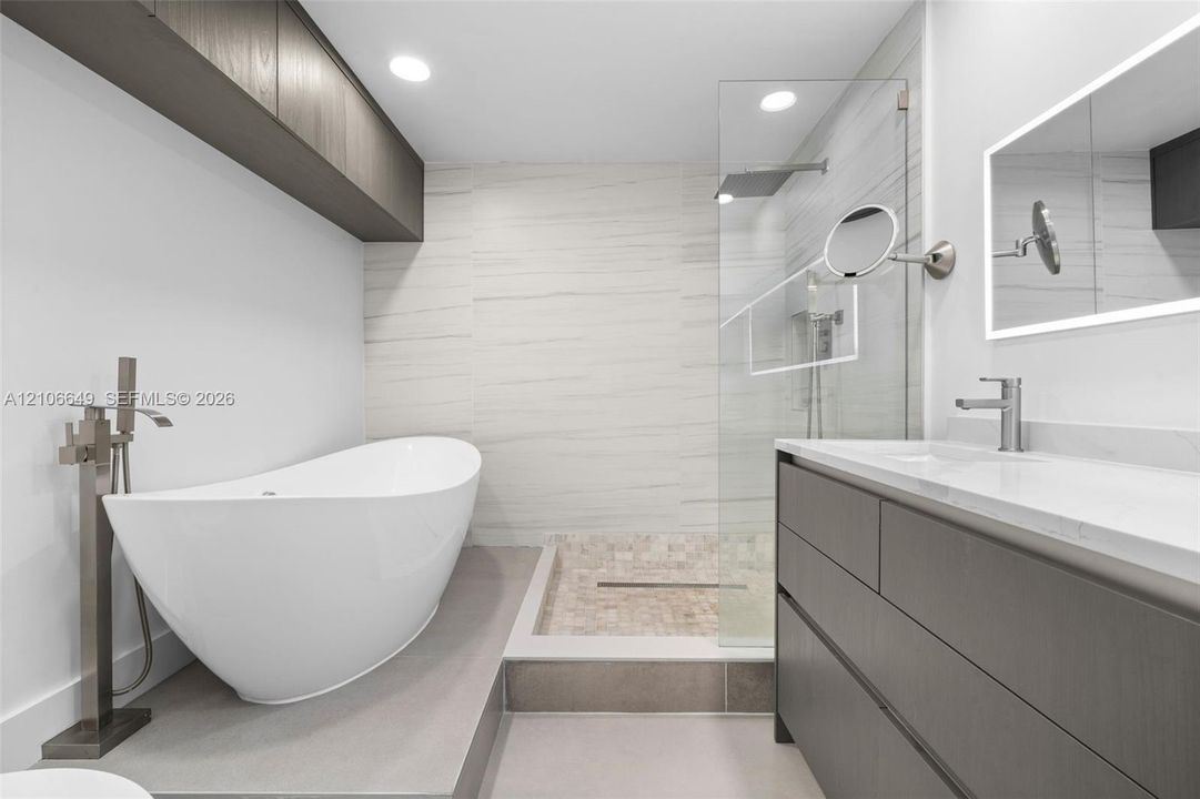 Freestanding soaking tub with floor-mounted faucet, walk-in glass-enclosed shower featuring mosaic stone flooring, dark wood-finish cabinetry, illuminated wall-mounted mirror, and recessed lighting