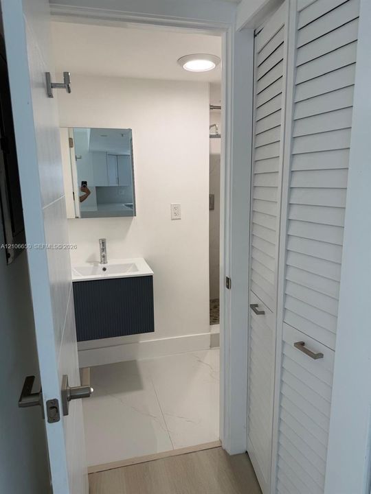 Floating vanity with a rectangular sink, wall-mounted mirror, recessed ceiling lighting, light-colored tile flooring, and white louvered-style closet doors